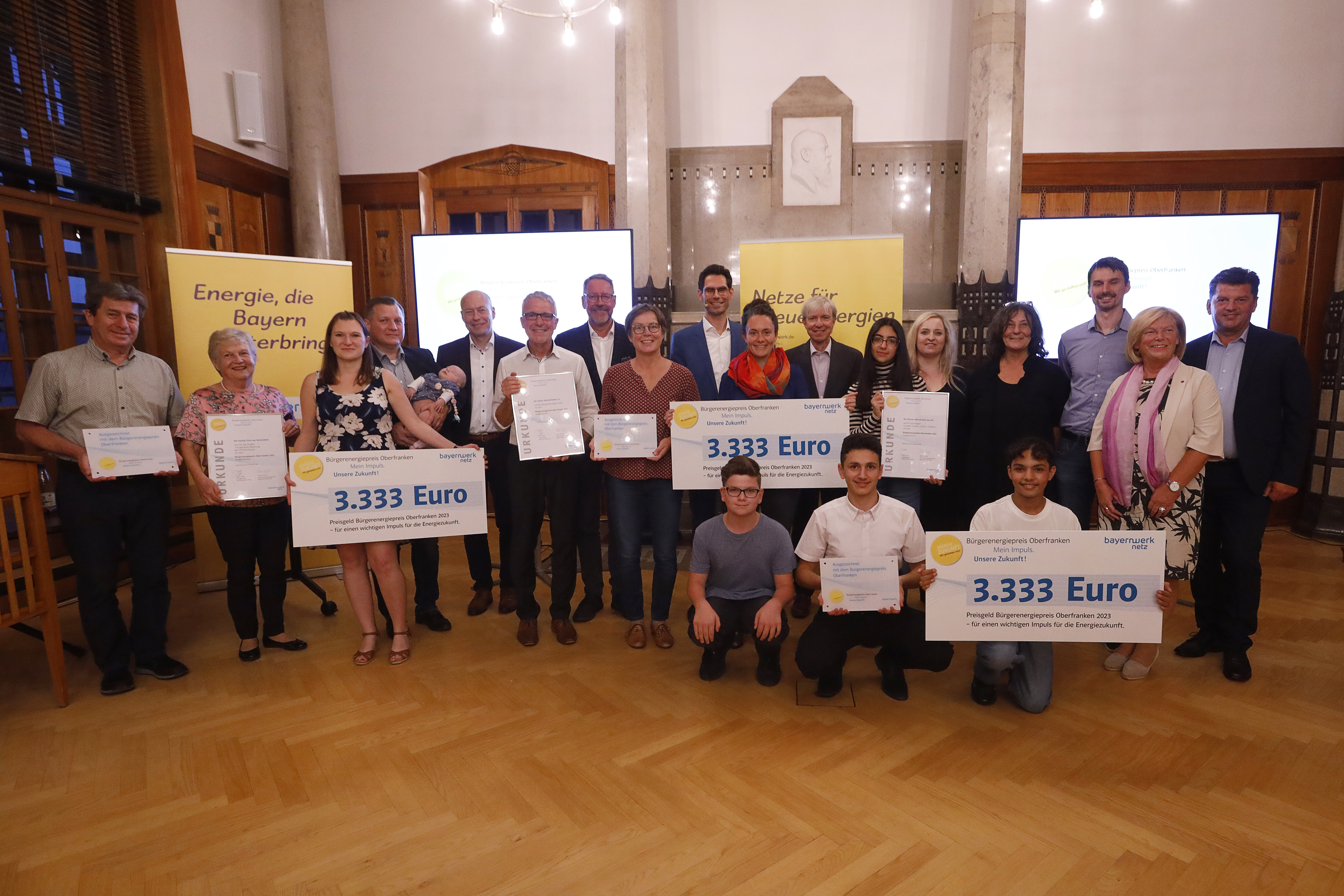 Die oberfränkischen Preisträger des Bürgerenergiepreises 2023. Gruppenfoto von mehreren Männern und Frauen von denen drei Personen ein Schild hochhalten auf dem steht "3.333 Euro".