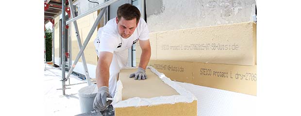Ein weiß gekleideter, dunkelhaariger Handwerker streicht mit einem Spatel weiße Paste auf einen kniehohen, länglichen Holzfaser-Klotz.