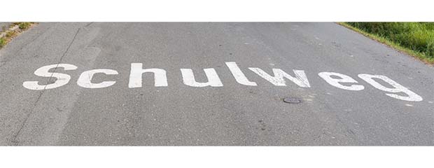 Das Wort "Schulweg" in weißen Buchstaben auf grauen Straßenasphalt geschrieben.