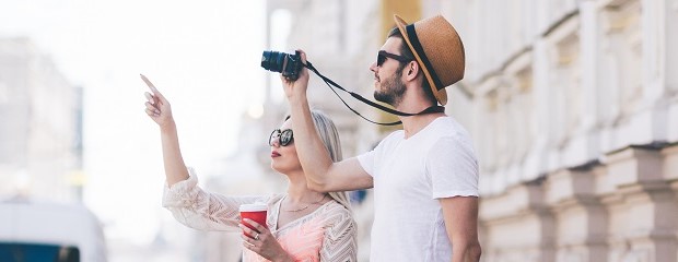 Ein junger Mann mit Sonnenbrille und Strohhut und eine junge, blonde Frau sehen sich ein Gebäude an. Der Mann hält eine Kamera hoch.