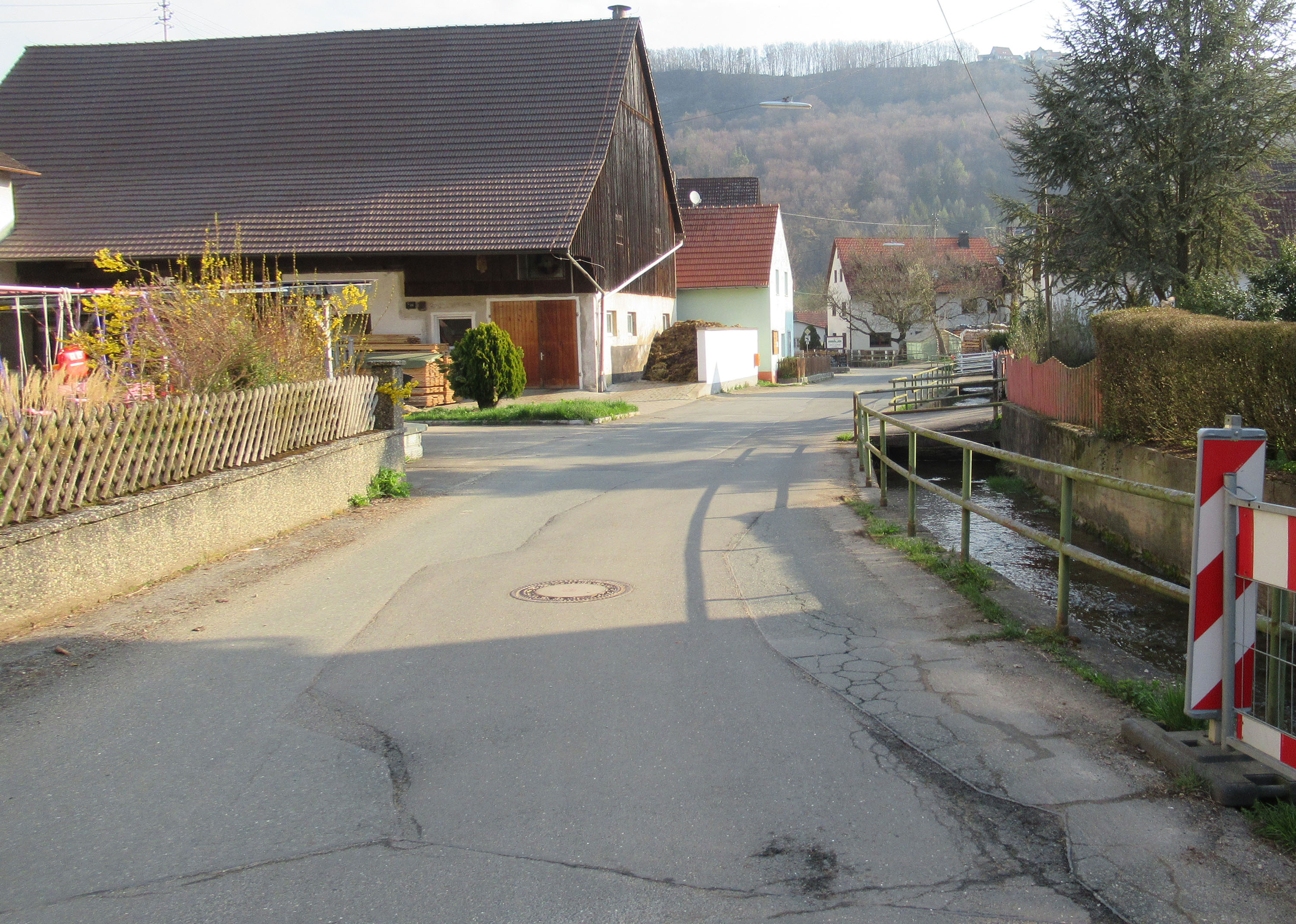 Blick aus Richtung Hundshaupten in die Ortsmitte von Oberzaunsbach mit schlechtem Fahrbahnzustand und erneuerungsbedürftiger Bachmauer und aufgesetztem Geländer.