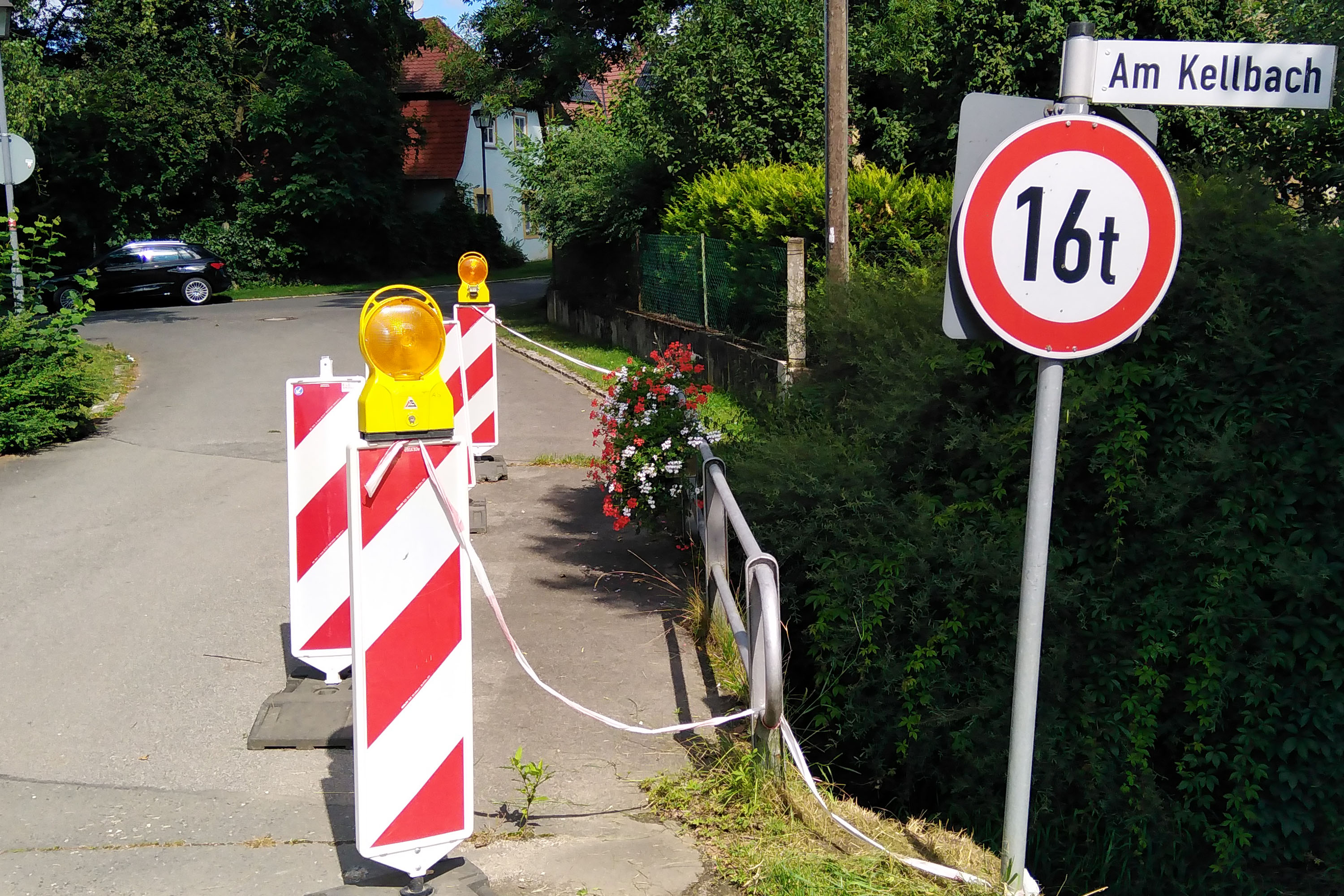 Vorhandene Situation an der Kellbachbrücke. Eine Brücke über einen kleinen Bach ist mit Pollern abgesperrt, weil der Straßenbelag beschädigt und rissig ist.
