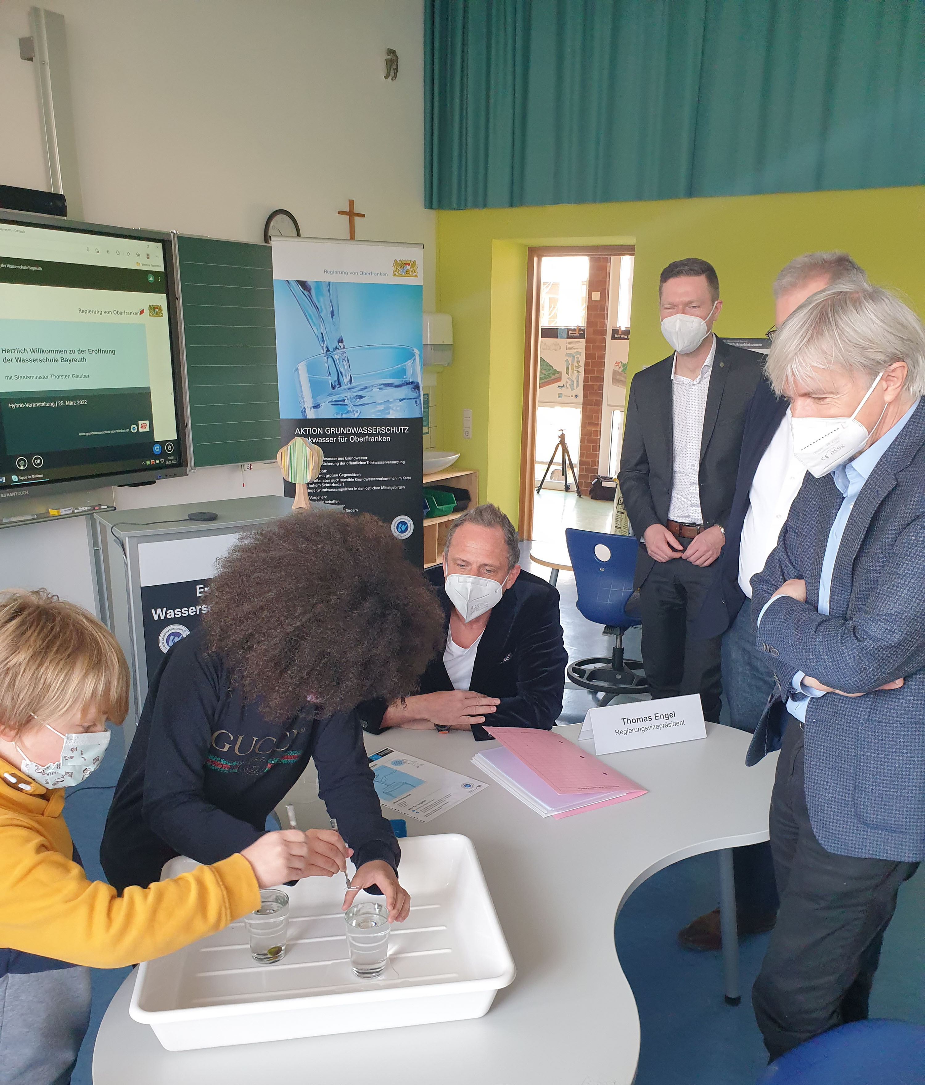 Staatsminister Thorsten Glauber (Mitte), Landrat Florian Wiedemann (3. von rechts) und Regierungsvizepräsident Thomas Engel (rechts) beobachten nach Eröffnung der neuen Wasserschule Bayreuth in der Grundschule Laineck interessiert die ersten Experimente.