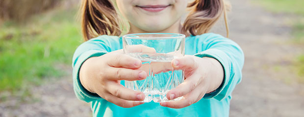 Ein lächelndes Mädchen mit türkisem Pulli hält vor sich ein Glas mit Leitungswasser hoch