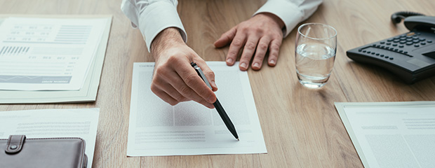 Formulare und Merkblätter für angehende Rechtsreferendare Symbolfoto: Blick auf einen Schreibtisch mit Formularen und Telefon, während eine männliche Hand auf einem Blatt etwas schreibt.