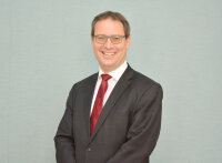 Regierungspräsident Florian Luderschmid Man sieht den Oberkörper eines Mannes mittleren Alters mit braunen kurzen Haaren und einer grauen Brille, der in die Kamera lächelt. Der Mann trägt einen dunkelgrauen Anzug und eine weinrote Krawatte auf weißem Hemd.
